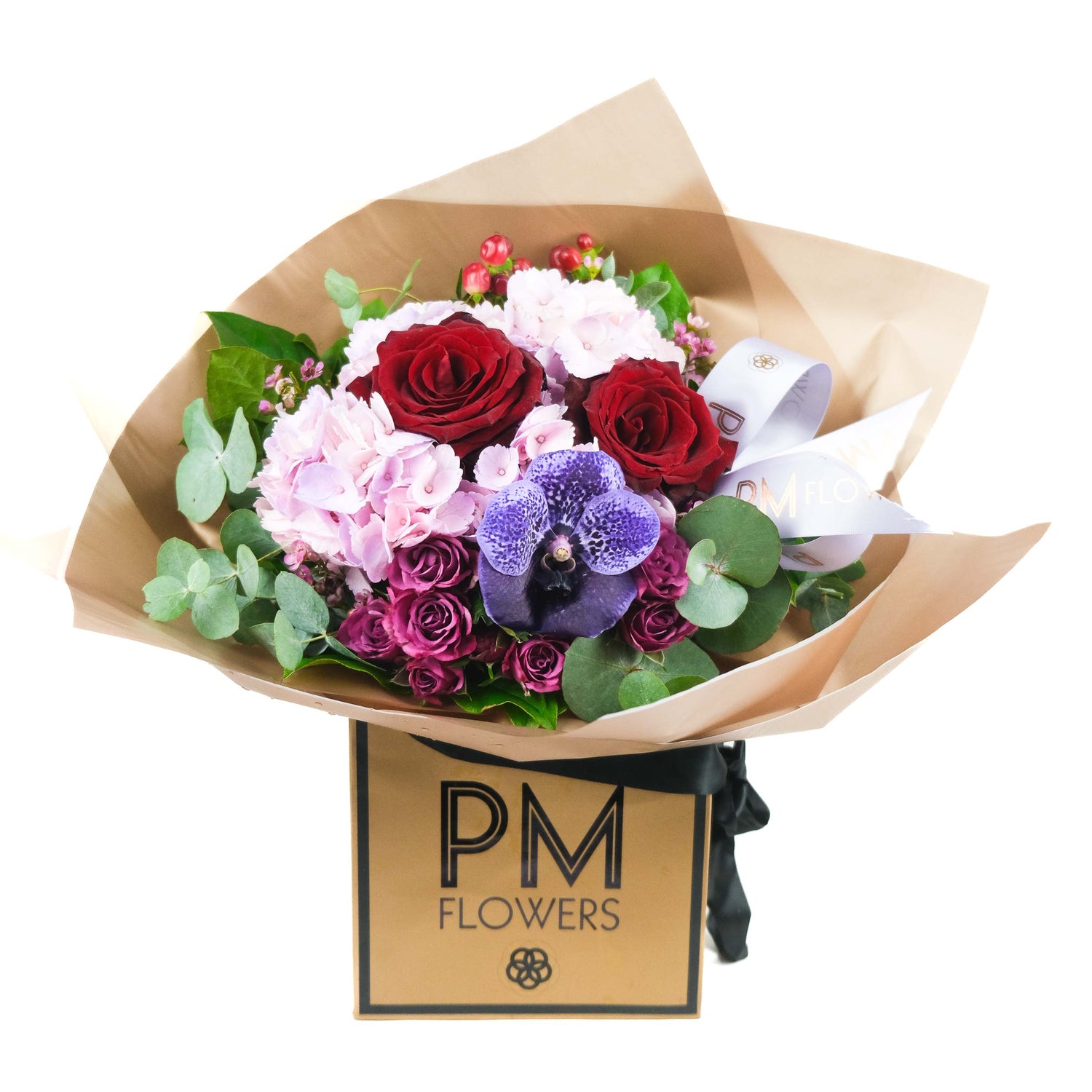 A bouquet of flowers consisting of pink hydrangea, red roses, red hypericum, pink waxflower, and purple spray roses, wrapped in Gold cello with a ribbon, presented in a card box with &