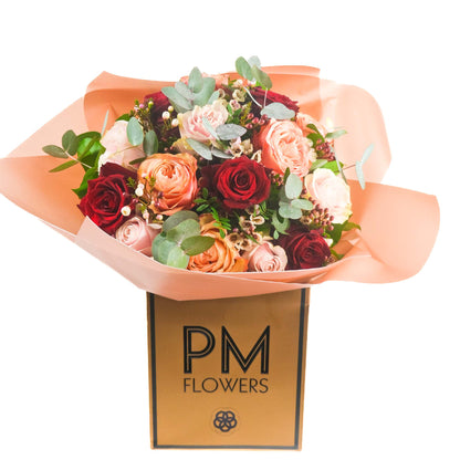 A bouquet of roses in shades of red, pink, and peach, with white waxflowers and green foliage, wrapped in paper with the &