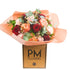 A bouquet of roses in shades of red, pink, and peach, with white waxflowers and green foliage, wrapped in paper with the &