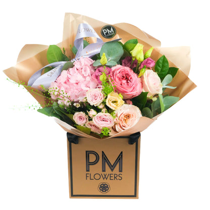 A bouquet of pink and peach flowers including roses and hydrangea, wrapped in a brown paper with a label that reads &