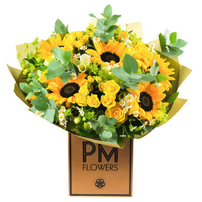 A vibrant bouquet of sunflowers, yellow spray roses, and various other flowers with green foliage, presented in a brown paper wrap with &